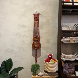 Könnte beinhalten: Eine braune Ukulele hängt in einem Makramee-Pflanzenaufhänger an einer strukturierten grauen Wand. Ein kleiner Holztisch trägt einen geflochtenen Korb mit einem roten Stofftier und einer kleinen Pflanze. Ein dunkles Holzregal ist im Hintergrund zu sehen.