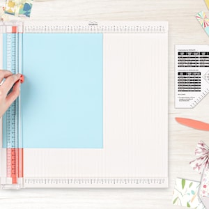 May include: A white paper trimmer with a ruler and a blue cutting mat. The trimmer is on a wooden surface with various craft supplies.