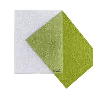 May include: Two sheets of clear plastic embossing stencils with a snowflake pattern. The top sheet is translucent and the bottom sheet is opaque. The sheets are layered on top of each other and the top sheet is folded over to show the pattern on the bottom sheet. The bottom sheet has the word "birdbirra" printed on it.