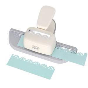May include: A cream-colored paper craft punch with a gray base and a lever. The punch is designed to create a decorative floral edge on paper. A light blue paper strip is being punched, and another cut strip is visible.