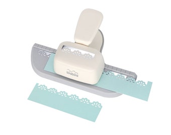 May include: A cream-coloured paper craft punch with a grey base and a lever. The punch is designed to create a decorative floral edge on paper. A light blue paper strip is being punched, and another cut strip is visible.