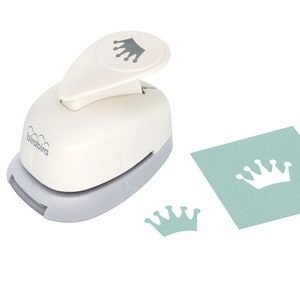 May include: White paper punch with a gray base. The punch is shaped like a crown and has a gray crown icon on the handle. The punch is shown next to a light blue piece of paper with a white crown cut out of it. There is also a white crown cut out of the paper on the table.