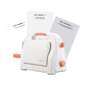 May include: A white die-cutting machine with a pink handle and a gray cutting mat. The machine has a dial for adjusting the cutting depth. The machine is labeled "Cricut" and the mat is labeled "Cutting Mat 12" x 12".