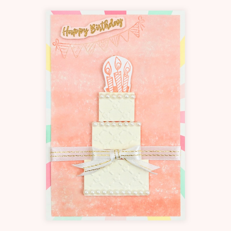 May include: A pink card with a white cake with candles on top. The cake is decorated with white pearls. The card says "Happy Birthday" in gold letters.
