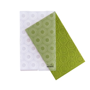 May include: A set of two clear plastic embossing mats with a repeating pattern of stylized sunbursts. The mats are layered on top of each other, with the top mat showing a white outline of the sunbursts and the bottom mat showing a green embossed version of the pattern.