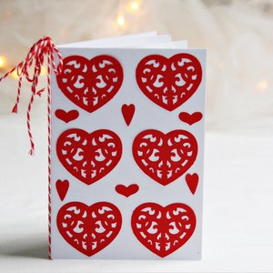 May include: A white card with red paper hearts cut out in a decorative pattern. The hearts are arranged in a grid pattern with smaller hearts in between the larger hearts.