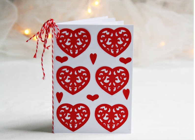 May include: A white card with red paper hearts cut out in a decorative pattern. The hearts are arranged in a grid pattern with smaller hearts in between the larger hearts.
