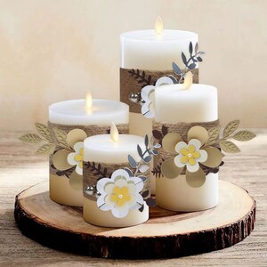 May include: Four white pillar candles with a rustic, farmhouse theme. Each candle is decorated with a burlap ribbon, a small flower, and a sprig of leaves. The candles are arranged on a wooden slice.