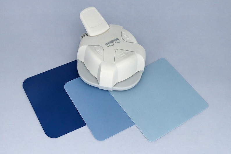 May include: A white paper punch with a blue, light blue, and dark blue paper samples. The punch has a lever and a circular cutting area. The paper samples are squares.