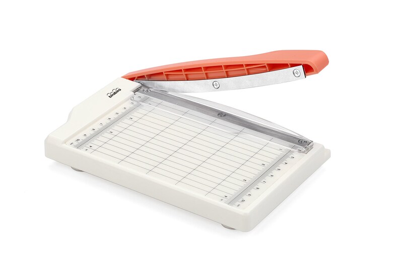 May include: A white paper trimmer with a clear cutting guide and an orange handle. The trimmer has a ruler on the side and a grid pattern on the cutting surface.