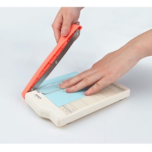 May include: A white paper trimmer with a red handle and a blue cutting mat. The handle is being pressed down to cut a piece of paper.