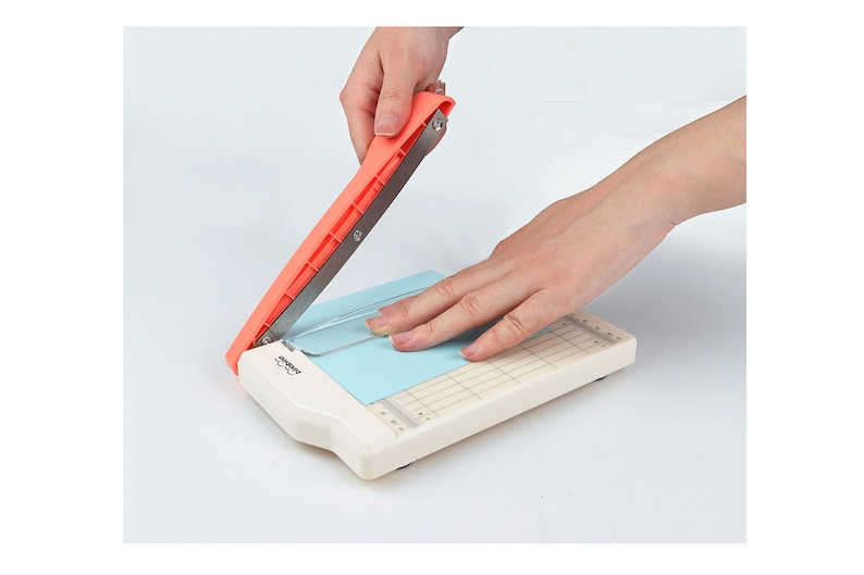 May include: A white paper trimmer with a red handle and a blue cutting mat. The handle is being pressed down to cut a piece of paper.