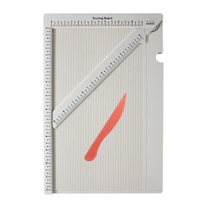 May include: A white scoring board with vertical grooves and inch measurements. A white ruler and a coral-colored scoring tool are included. The words "Scoring Board" are printed on the top. The brand name "biribiro" is also visible.