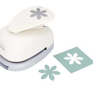 May include: White paper punch with a gray flower design on the top. The punch is used to create a flower shape from paper. Two paper flowers are shown, one in front of the punch and one on a blue piece of paper.