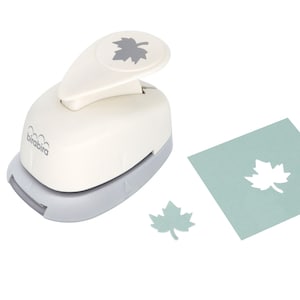 Op de afbeelding: Een witte en grijze papierpons met een bladerenontwerp. De pons staat op een witte achtergrond met een groene bladeren-uitsparing en een kleinere bladeren-uitsparing.