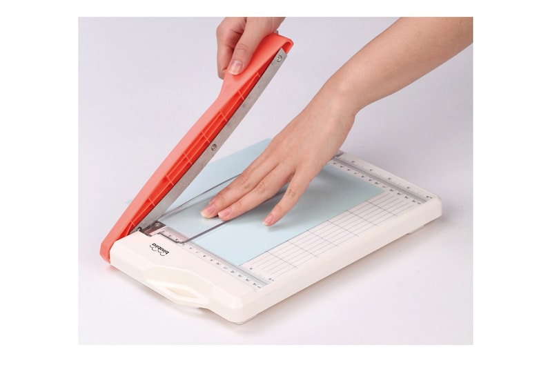 Puede incluir: Un cortador de papel blanco con un mango rojo y una cuchilla plateada. El cortador tiene una regla en el lateral y un patr&oacute;n de cuadr&iacute;cula en la superficie de corte. Una mano sostiene el mango y presiona la cuchilla hacia abajo para cortar un trozo de papel azul.