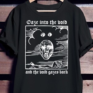 Puede incluir: Camiseta negra con un diseño gráfico blanco. El diseño presenta una cara estilizada con la boca abierta, sobre un fondo nublado. El texto "Gaze into the void" está encima de la imagen, y "and the void gazes back" debajo.