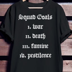 Könnte beinhalten: Schwarzes T-Shirt mit dem Text "Squad Goals" und einer Liste in gotischer Schrift: I. Krieg, II. Tod, III. Hungersnot, IV. Pestilenz. Das T-Shirt hängt an einem Kleiderbügel.