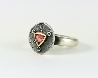 May include: A silver ring with a round, textured bezel set with a triangular pink gemstone. The bezel is accented with small gold circles.