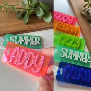 Peut inclure: Plusieurs étiquettes nominatives en acrylique coloré, aux couleurs vives comme le rose, l'orange, le vert et le bleu. Chaque étiquette affiche un nom ou un mot en couleur contrastante, comme "Addy", "Elijah", "Ozzy", "Summer" et "Oluwa".