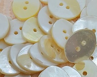 Könnte beinhalten: Ein Haufen ovaler Knöpfe in Weiß und Creme mit jeweils zwei Löchern. Die Knöpfe sind aus perlmuttartigem Material gefertigt und haben eine glatte, glänzende Oberfläche.