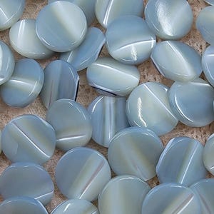 Peut inclure: Un ensemble de perles œil de chat bleu pâle et blanches. Chaque perle ronde présente une rayure blanche diagonale, créant un effet visuel unique. Les perles sont dispersées sur une surface texturée de couleur claire.