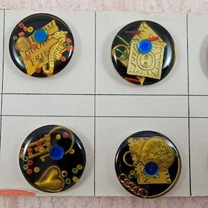 May include: Eight round black buttons with gold and blue accents. Each button features a different design with a variety of images including a heart, a crown, a key, a star, a seashell, a coin, and a piece of paper with the words "Good Friend".