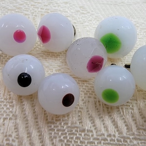 May include: A collection of ten white glass eyes with various colored pupils. The pupils are in different shapes and colors, including blue, green, pink, and black.