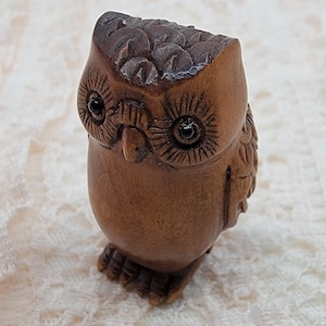 May include: A small, intricately carved wooden owl figurine. The owl has detailed feather and eye markings, with a rounded body and a slightly tilted head. The wood has a warm, brown tone, and the owl stands on small feet.