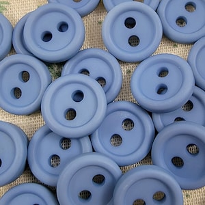 Puede incluir: Una colección de 20 botones de plástico azul, cada uno con dos agujeros. Los botones están dispuestos en un patrón aleatorio sobre un fondo de tela marrón claro.