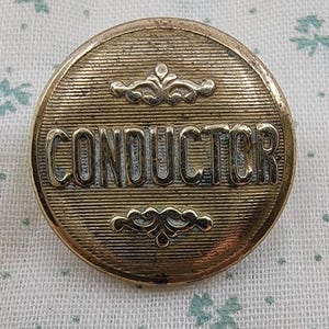 May include: A circular gold-toned metal button featuring the word CONDUCTOR embossed in capital letters across the center. The button has a fine horizontal line texture and decorative scrollwork motifs positioned above and below the text.