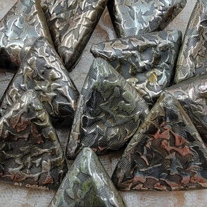 May include: A cluster of silver-toned, triangular decorative pieces with a textured, embossed pattern. The pieces are arranged together, highlighting their metallic sheen and intricate design. The background is a light, patterned fabric.