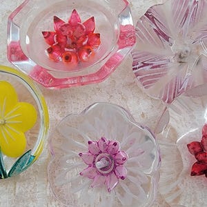 May include: A collection of colorful, floral-themed coasters. The coasters feature various flower designs in shades of yellow, pink, and white, encased in clear resin. The shapes include round, octagonal, and flower-shaped designs.