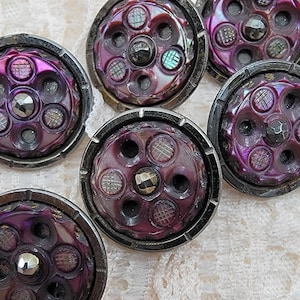 Può includere: Un set di cinque bottoni circolari con finitura viola metallizzato e bordo grigio scuro. Ogni bottone presenta una gemma centrale sfaccettata circondata da cinque rientranze circolari, quattro delle quali contengono piccoli motivi a griglia.