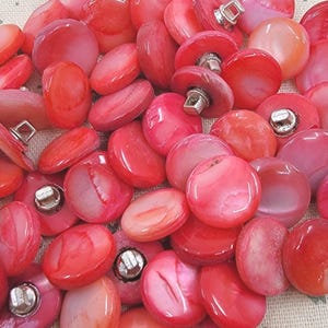 May include: A collection of red and pink shell buttons with silver metal shanks. The buttons vary in size and shape, with a mix of round and oval designs. The buttons are scattered on a light-colored fabric with a subtle floral pattern.