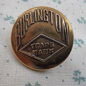 May include: A round, metallic gold-toned button featuring the embossed text BURLINGTON in an arc at the top. Below the text, a diamond-shaped border encloses the words TRADE MARK in raised lettering, creating a distinct, vintage-style design.