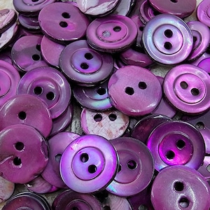 Può includere: Un grande mucchio di bottoni di conchiglia iridescente viola con due fori ciascuno. I bottoni sono disposti in modo casuale e sono tutti della stessa dimensione.