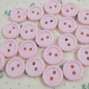 Puede incluir: Una colección de 20 botones de plástico rosa con dos agujeros cada uno. Los botones están dispuestos en un patrón circular sobre un fondo de tela rosa claro.