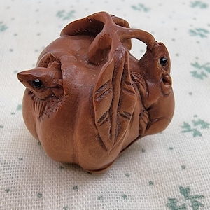 May include: A small, brown, hand-carved wooden netsuke featuring a detailed pumpkin or fruit shape. The carving includes intricate leaves and a small animal figure with dark, inlaid eyes integrated into the organic, rounded form of the object.
