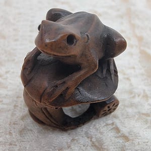 May include: A dark brown carved wooden ring featuring a detailed frog perched atop a mushroom cap. The band of the ring is sculpted with organic textures and a small circular floral motif on the side, showcasing intricate craftsmanship.