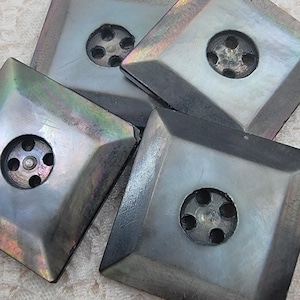 May include: Four square buttons with a dark, iridescent, metallic-like finish. Each button features a beveled edge leading to a central circular depression containing four small holes for attachment, displaying subtle rainbow-toned reflections.