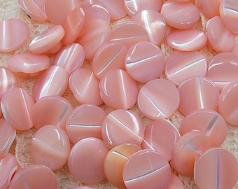 May include: A collection of light pink, oval buttons with a pearlescent finish. Each button features a white, diagonal stripe across the middle. The buttons are scattered on a textured, off-white surface.
