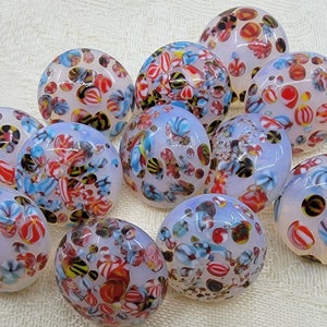 May include: A collection of 12 round, white glass buttons with colorful, intricate patterns. The buttons have a translucent, milky white appearance and feature a variety of colors, including red, blue, and black.