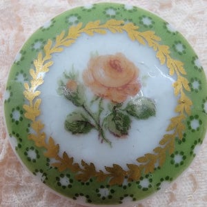May include: A round, white porcelain button with a green and gold design. The button features a single yellow rose with green leaves and a gold leaf border.