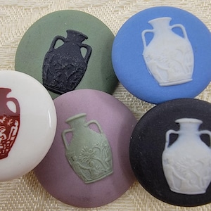 Puede incluir: Un conjunto de botones redondos decorativos en varios colores, cada uno con un diseño en relieve de un jarrón clásico. Los colores incluyen blanco, negro, azul, verde y lavanda. Los diseños de los jarrones varían en color y detalle.