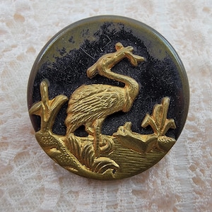 May include: Round button with a gold-colored crane design on a black and green background. The crane is standing in a marshy area with gold-colored plants. The button has a shiny finish.