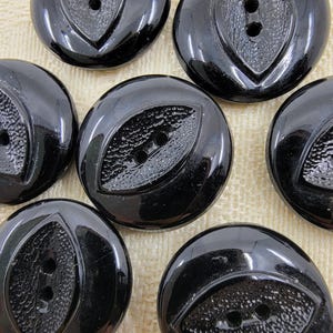 May include: A collection of glossy black buttons. Each button is round with a textured, eye-shaped design in the center, featuring two small holes for sewing. The buttons have a smooth, reflective surface.