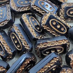 May include: A collection of black rectangular beads with gold-colored filigree and oval-shaped accents. The beads have a glossy finish and are arranged closely together, showcasing their intricate design.