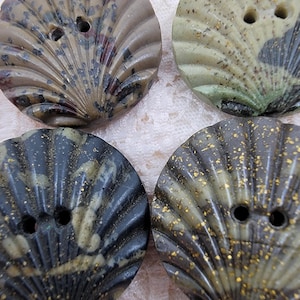 May include: Four seashell-shaped buttons in shades of brown, green, and gray with speckled and gold accents. Each button has two holes for sewing. The buttons have a ridged, fan-like design.