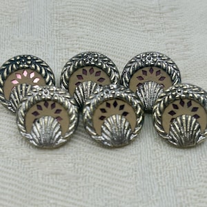 Può includere: Sei bottoni vintage color argento con un design decorativo. Ogni bottone presenta una forma a conchiglia smerlata, un bordo floreale e un centro beige con un accento rosa. I bottoni sono disposti su una superficie strutturata bianco sporco.